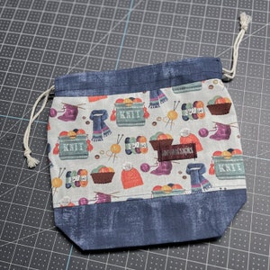 Sock knitting bag with knitting fabric print, drawstring closure and inner dividing zippered pocket and snap tabs to keep yarn from tangling