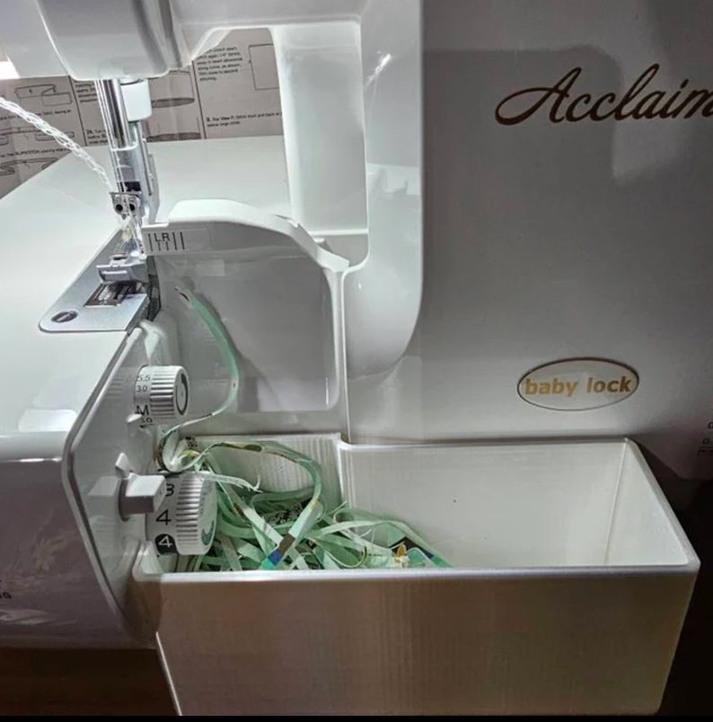 May include: White Baby Lock Acclaim serger with a clear plastic bin filled with green and blue thread scraps. The serger has a white dial with numbers 3, 4, 5, 6, 7, 8, 9, 10, and a smaller white dial with numbers 1, 2, 3, 4, 5.