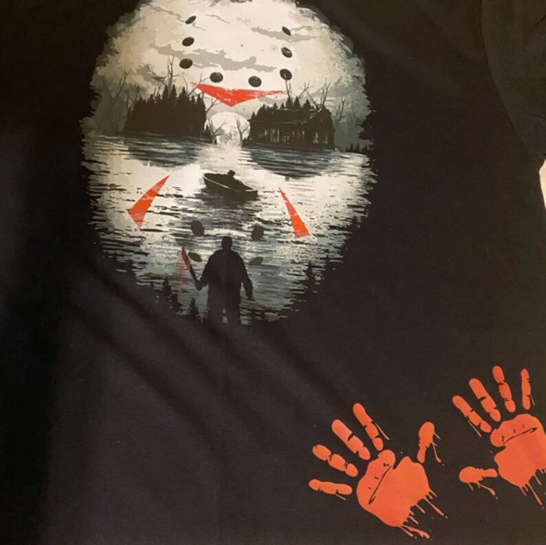 May include: Black t-shirt with a white and red graphic of a hockey mask reflecting in water. The graphic also includes a silhouette of a person holding a knife and a boat. There are two red handprints on the bottom of the shirt.