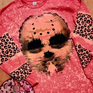 May include: A pink long-sleeved shirt with a bleach-splattered design featuring a silhouette of a hockey mask and a figure in a boat. The sleeves have a leopard print pattern.