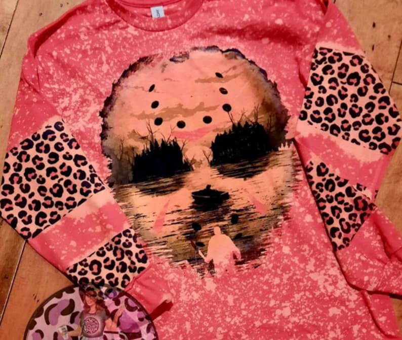 May include: A pink long-sleeved shirt with a bleach-splattered design featuring a silhouette of a hockey mask and a figure in a boat. The sleeves have a leopard print pattern.
