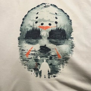 May include: A white t-shirt with a graphic print of a hockey mask reflected in a lake. The mask is partially obscured by trees and a boat in the water. A figure with a knife stands in the foreground.