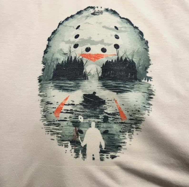 May include: A white t-shirt with a graphic print of a hockey mask reflected in a lake. The mask is partially obscured by trees and a boat in the water. A figure with a knife stands in the foreground.