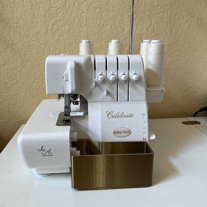 May include: White Baby Lock Celebrate serger sewing machine with a gold-colored accessory tray. The machine has a clear plastic spool holder with four spools of white thread. The machine has a Jet Air threading system.
