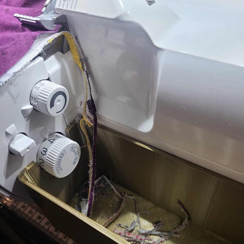 May include: Close-up of a white sewing machine with a gold-colored compartment. The compartment is filled with scraps of fabric and thread. The machine has two dials labeled "Standard" and "Rolled Hem".