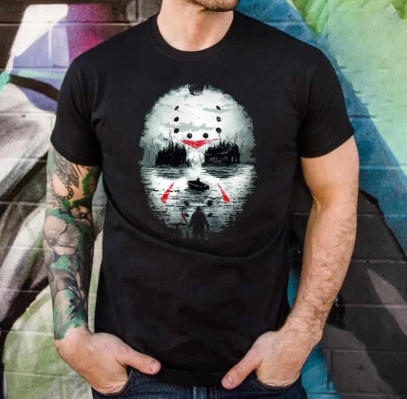 May include: A black t-shirt with a white and red graphic print of a hockey mask with a forest scene in the background.