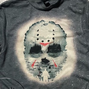 May include: A gray t-shirt with a white bleach design of a hockey mask with a silhouette of a figure standing in front of a cabin in the woods. The mask is partially submerged in water.