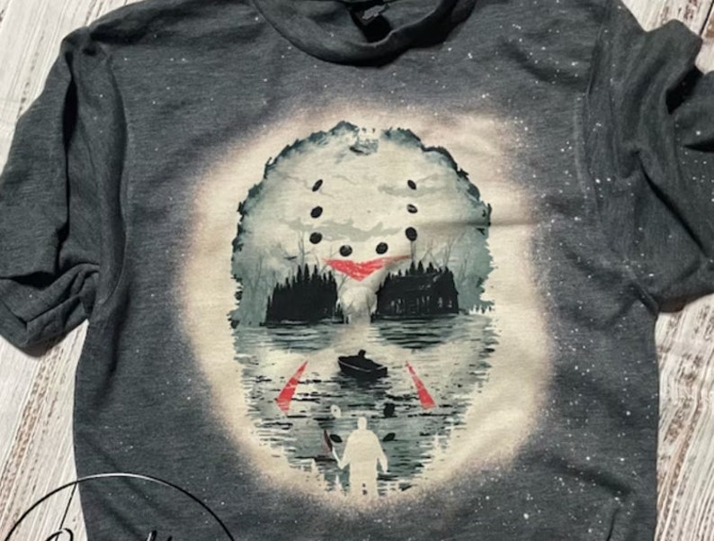 May include: A gray t-shirt with a white bleach design of a hockey mask with a silhouette of a figure standing in front of a cabin in the woods. The mask is partially submerged in water.