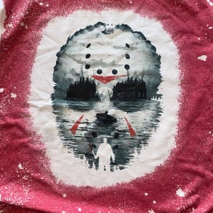 May include: A white t-shirt with a red splatter paint design. The design features a hockey mask with a lake scene in the background. A figure holding a machete is standing in the water.