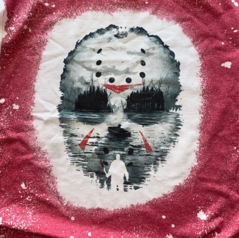 May include: A white t-shirt with a red splatter paint design. The design features a hockey mask with a lake scene in the background. A figure holding a machete is standing in the water.