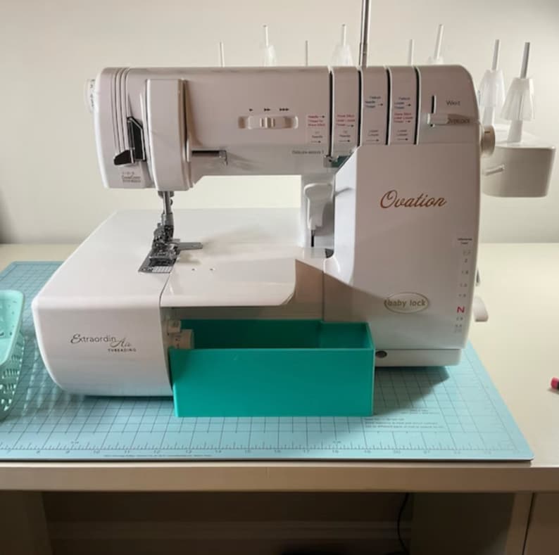 May include: White Baby Lock Ovation serger with a turquoise accessory drawer. The serger has a clear plastic thread guide and a white plastic needle plate. The serger is on a blue and white gridded cutting mat.