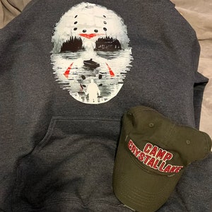 May include: A dark gray hooded sweatshirt with a white graphic of a hockey mask with a red slash across the eyes. The mask is superimposed over a scene of a house and a boat on a lake. A green baseball cap with the text "Camp Crystal Lake" in red is on the sweatshirt.