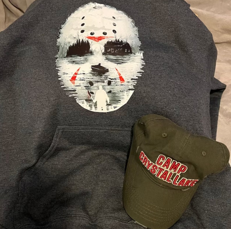 May include: A dark gray hooded sweatshirt with a white graphic of a hockey mask with a red slash across the eyes. The mask is superimposed over a scene of a house and a boat on a lake. A green baseball cap with the text "Camp Crystal Lake" in red is on the sweatshirt.
