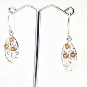 May include: Silver dangle earrings with a leaf design and multiple small, round, yellow gemstones.