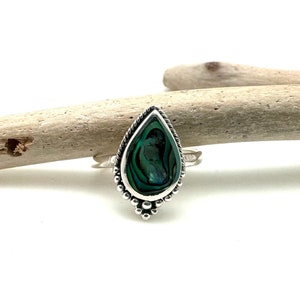 K&ouml;nnte beinhalten: Ein silberner Ring mit einem tropfenf&ouml;rmigen Einlegeteil aus Abalone-Schale. Der Ring hat einen perlenbesetzten Rand und liegt auf einer Holzoberfl&auml;che.