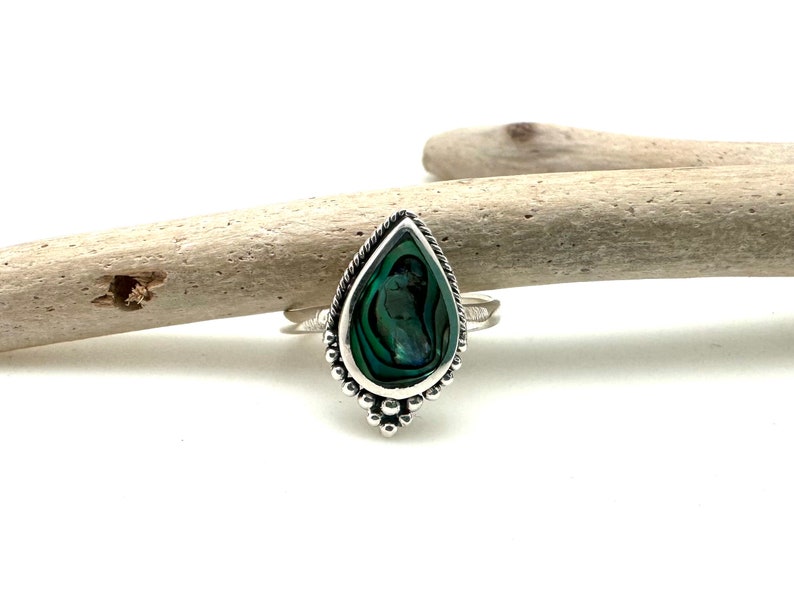 K&ouml;nnte beinhalten: Ein silberner Ring mit einem tropfenf&ouml;rmigen Einlegeteil aus Abalone-Schale. Der Ring hat einen perlenbesetzten Rand und liegt auf einer Holzoberfl&auml;che.