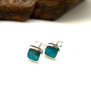 May include: A pair of stud earrings featuring square-shaped turquoise stones set in polished silver frames. Each earring has a smooth, rounded square profile with a vibrant blue-green stone surface, showcasing natural dark matrix veining throughout.