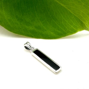 May include: A rectangular pendant with a polished silver setting and a glossy black inlay. The pendant is suspended from a small silver loop. The background is white with a green leaf.