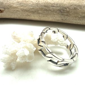 May include: A silver ring featuring a chain link design. The ring is made of polished metal and has a textured surface. The ring is displayed on a white surface with white coral and driftwood in the background.