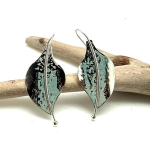 May include: A pair of silver-toned earrings shaped like leaves with a textured, mottled surface featuring shades of light blue, dark blue, and metallic silver. Each earring has a thin, curved silver wire stem and a small ball at the bottom tip.