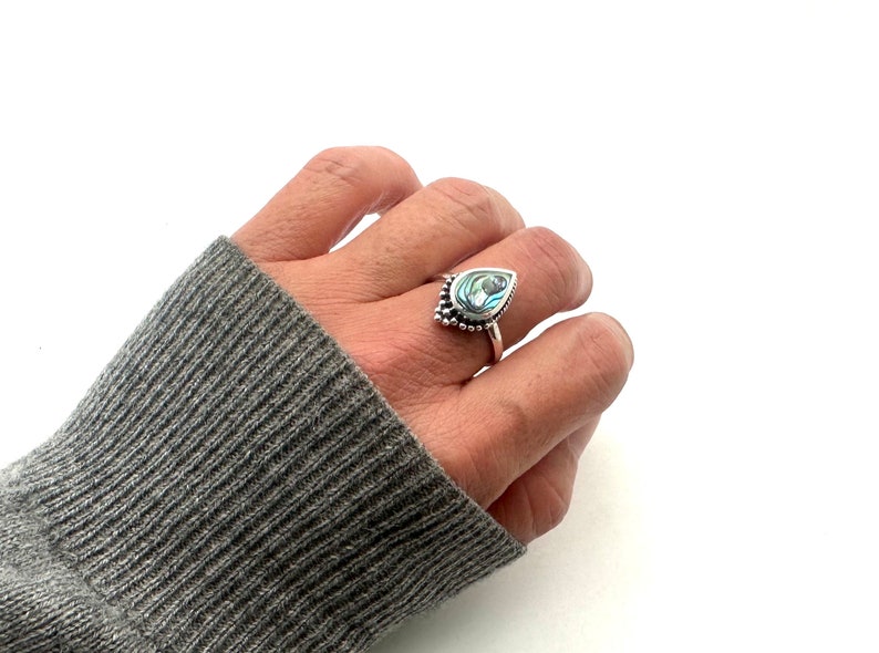 K&ouml;nnte beinhalten: Ein silberner Ring mit einem tropfenf&ouml;rmigen Abalone-Muschel-Stein. Der Ring hat ein Perlenmuster um den Stein.
