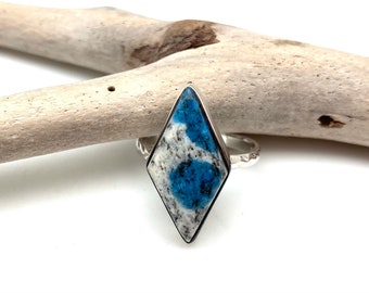 Peut inclure: Bague en argent ornée d'une grande pierre en forme de losange avec un motif bleu et blanc saisissant. La pierre est sertie dans une lunette en argent, et le corps de la bague présente un motif texturé. La bague est présentée sur un morceau de bois flotté.