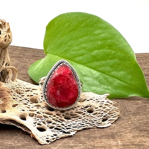 May include: A teardrop-shaped red gemstone ring with a silver setting. The ring is set on a piece of driftwood with a green leaf behind it.