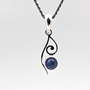 Op de afbeelding: Een zilveren ketting met een hanger met een blauwe edelsteen en een delicaat, zwierig ontwerp.