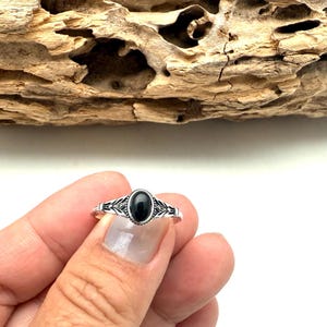 K&ouml;nnte beinhalten: Ein silberner Ring mit einem ovalen, schwarzen Stein. Der Ring hat ein detailliertes, dekoratives Design um den Stein und am Band. Der Ring wird vor einem St&uuml;ck Treibholz pr&auml;sentiert.