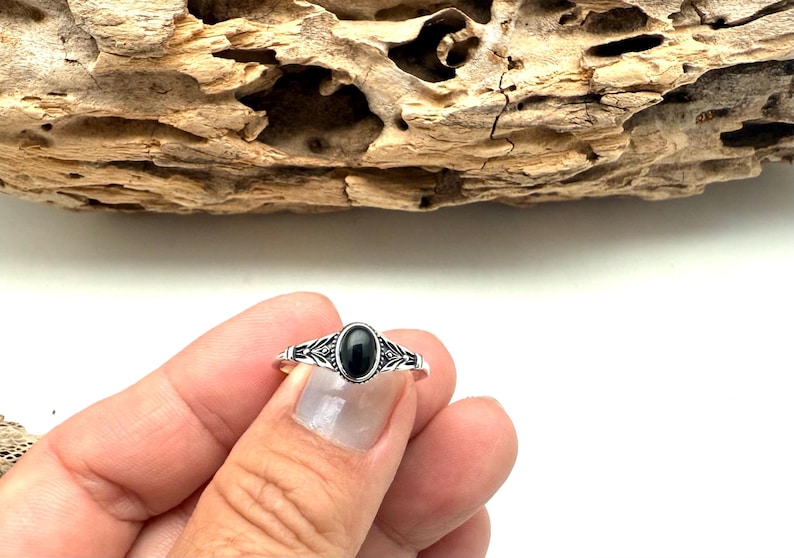 K&ouml;nnte beinhalten: Ein silberner Ring mit einem ovalen, schwarzen Stein. Der Ring hat ein detailliertes, dekoratives Design um den Stein und am Band. Der Ring wird vor einem St&uuml;ck Treibholz pr&auml;sentiert.