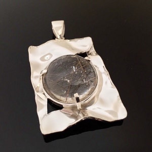 May include: A silver pendant with a large, round, dark gemstone with internal inclusions. The gemstone is set within a textured, rectangular silver frame. The pendant has a silver bail for a chain.