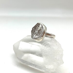 May include: A silver ring with a large, clear, faceted, rough-cut gemstone. The ring is set on a white crystal surface.