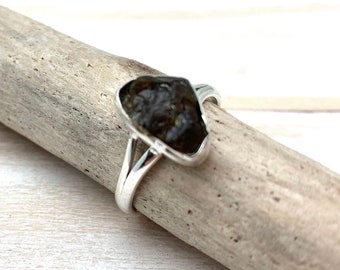 Puede incluir: Un anillo de plata con una piedra preciosa verde oscura engastada en una forma única y facetada. El anillo se muestra sobre un trozo de madera flotante.