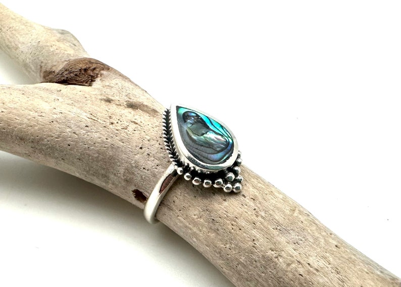 K&ouml;nnte beinhalten: Ein silberner Ring mit einem tropfenf&ouml;rmigen Einlegeteil aus Abalone-Muschel. Der Ring hat ein Perlenmuster um den Rand der Muschel.