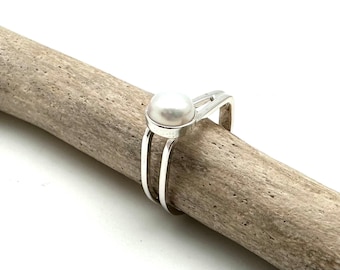 May include: A silver ring with a white pearl set in a modern design. The ring is on a piece of driftwood.