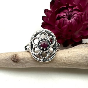 May include: A silver ring with a delicate floral design and a red gemstone in the center.
