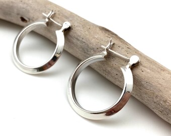 May include: A pair of silver hoop earrings with a simple, polished finish. The earrings are displayed on a piece of driftwood.