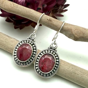 May include: A pair of sterling silver drop earrings with oval, faceted, red ruby gemstones. The earrings have a detailed, beaded border and are suspended from a simple hook. The background includes a wooden branch and a dark red flower.