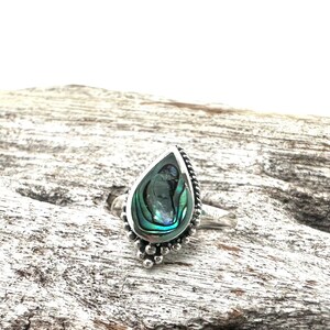 K&ouml;nnte beinhalten: Ein silberner Ring mit einem tropfenf&ouml;rmigen Einlegeteil aus Abalone-Muschel. Der Ring hat ein Perlenmuster um den Rand der Muschel.