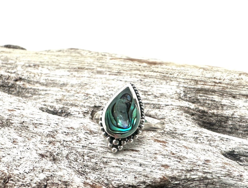 K&ouml;nnte beinhalten: Ein silberner Ring mit einem tropfenf&ouml;rmigen Einlegeteil aus Abalone-Muschel. Der Ring hat ein Perlenmuster um den Rand der Muschel.