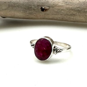 May include: A silver ring with a large, oval-shaped ruby gemstone. The ring has a simple band with small beads on each side of the stone.