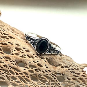 K&ouml;nnte beinhalten: Ein silberner Ring mit einem ovalen schwarzen Stein. Der Ring hat ein verziertes Design um den Stein und auf dem Band. Der Ring wird auf einem St&uuml;ck getrocknetem Holz mit hellbrauner Farbe und por&ouml;ser Textur pr&auml;sentiert.