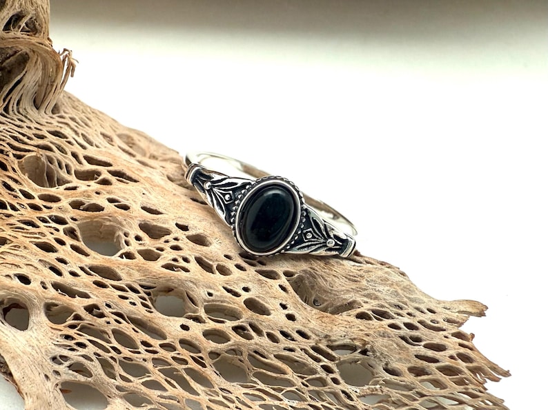 K&ouml;nnte beinhalten: Ein silberner Ring mit einem ovalen schwarzen Stein. Der Ring hat ein verziertes Design um den Stein und auf dem Band. Der Ring wird auf einem St&uuml;ck getrocknetem Holz mit hellbrauner Farbe und por&ouml;ser Textur pr&auml;sentiert.