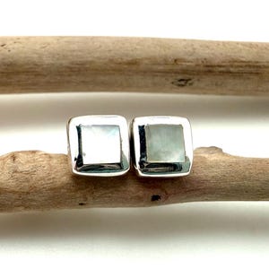 May include: A pair of square stud earrings featuring polished silver settings and inlaid iridescent square stones. The earrings have a smooth, rounded edge design and a reflective surface that catches the light, showcasing a subtle pearlescent sheen.