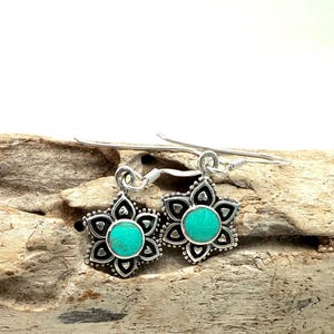Puede incluir: Un par de pendientes colgantes de plata con dijes de flores de color turquesa. Los pendientes tienen un diseño delicado y son perfectos para agregar un toque de color a cualquier atuendo.