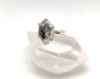 Può includere: Un anello d'argento con un cristallo trasparente, sfaccettato, di colore grigiastro incastonato al centro. Il cristallo ha la forma di un diamante.