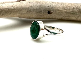 May include: A sterling silver ring featuring a large, oval-cut emerald gemstone. The emerald is a deep, vibrant green color and is set in a polished silver band. The ring is displayed on a white surface.