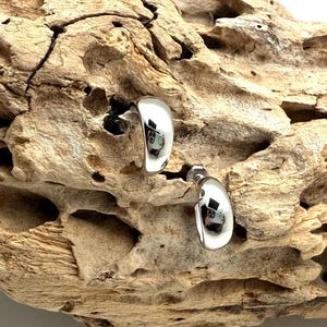 May include: A pair of polished silver earrings with a curved, teardrop design. The earrings are displayed against a textured, light brown piece of driftwood, creating a contrast between the smooth metal and the rough wood.