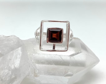 May include: A silver ring featuring a square, dark red gemstone set within a square frame. The ring's design includes a smaller square gemstone at the center, creating a layered effect. The ring is displayed on a clear, crystalline surface.