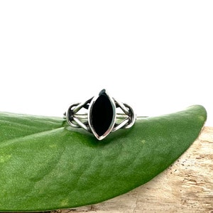 Może przedstawiać: Srebrny pierścionek z czarnym owalnym kamieniem szlachetnym osadzonym w delikatnym, skomplikowanym wzorze. Pierścionek leży na zielonym liściu.